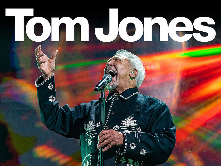 Tom Jones singt enthusiastisch am Mikrofon und die Daten seiner Tour 2024 werden eingeblendet.